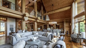 A spacious log cabin interior with a large L-shaped sectional sofa, a round coffee table, and a balcony with a railing.