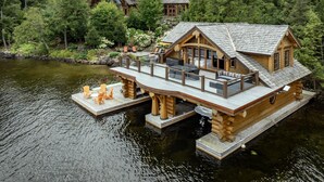 A log cabin with a deck extending over a lake, surrounded by dense forest.