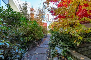 Ein schmaler Steinweg, gesäumt von üppigem Grün und Herbstlaub, führt zu einem Gebäude mit roter Fassade.