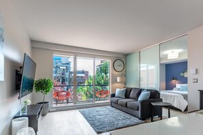 A modern living room with a large window, a flat-screen TV, a grey sofa, a small table, and a potted plant. - Sun-Drenched Sophistication (Victoria North)