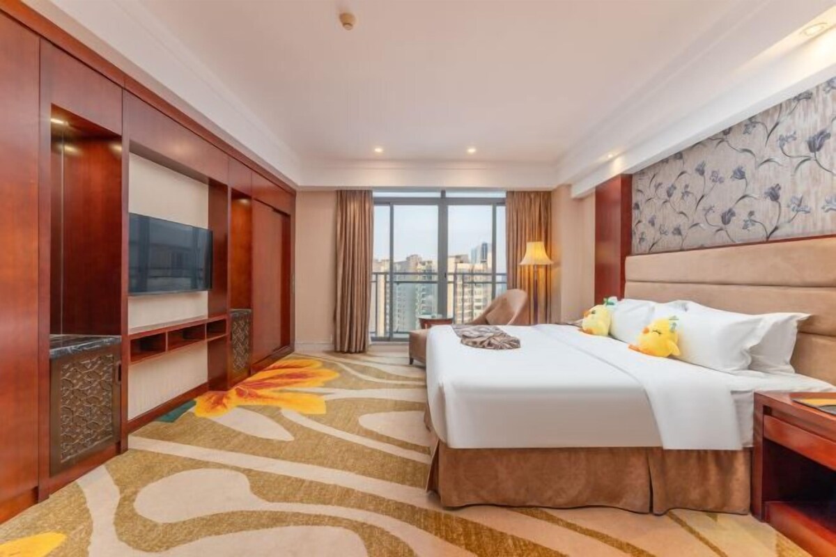 A hotel room with a large bed, a TV, a balcony with curtains, and a view of the city.