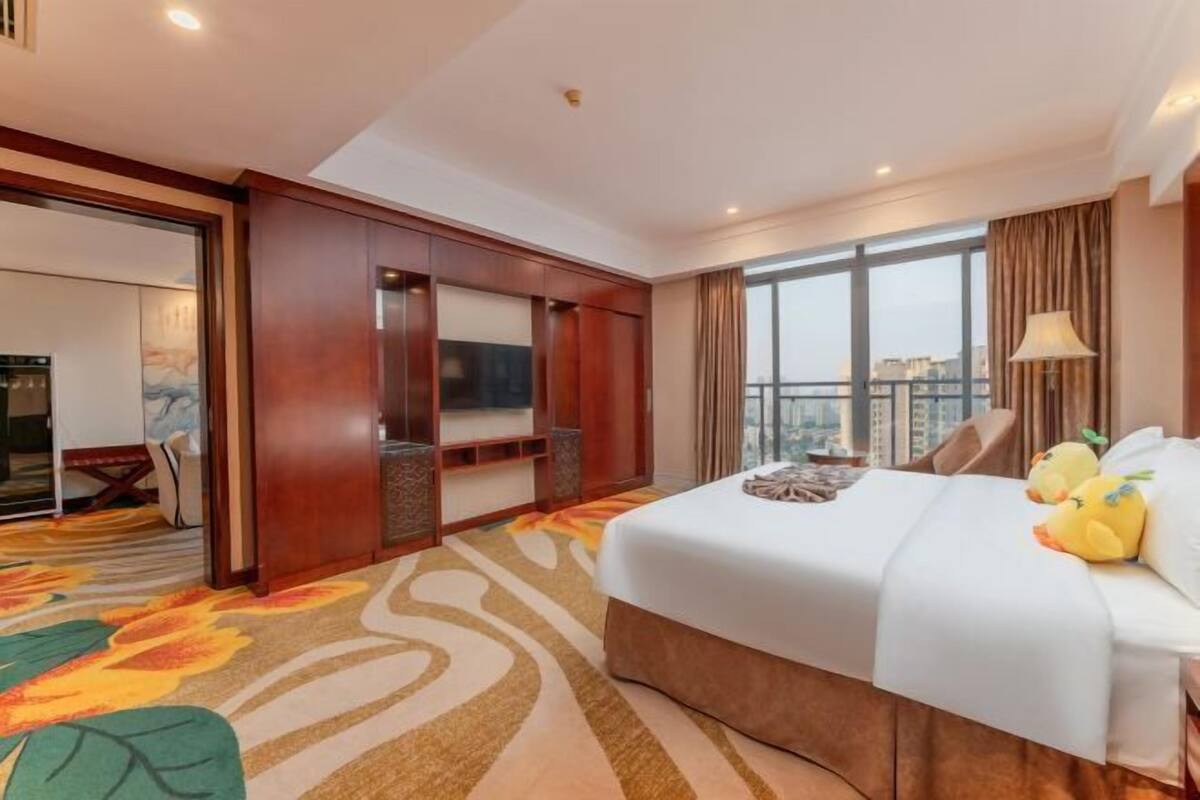 A hotel room with a large bed, a TV, a city view, and a wooden wardrobe.