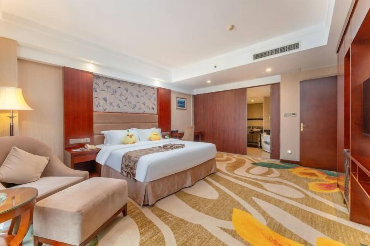 A hotel room with a bed, a desk, a chair, a bedside table, and a carpet.