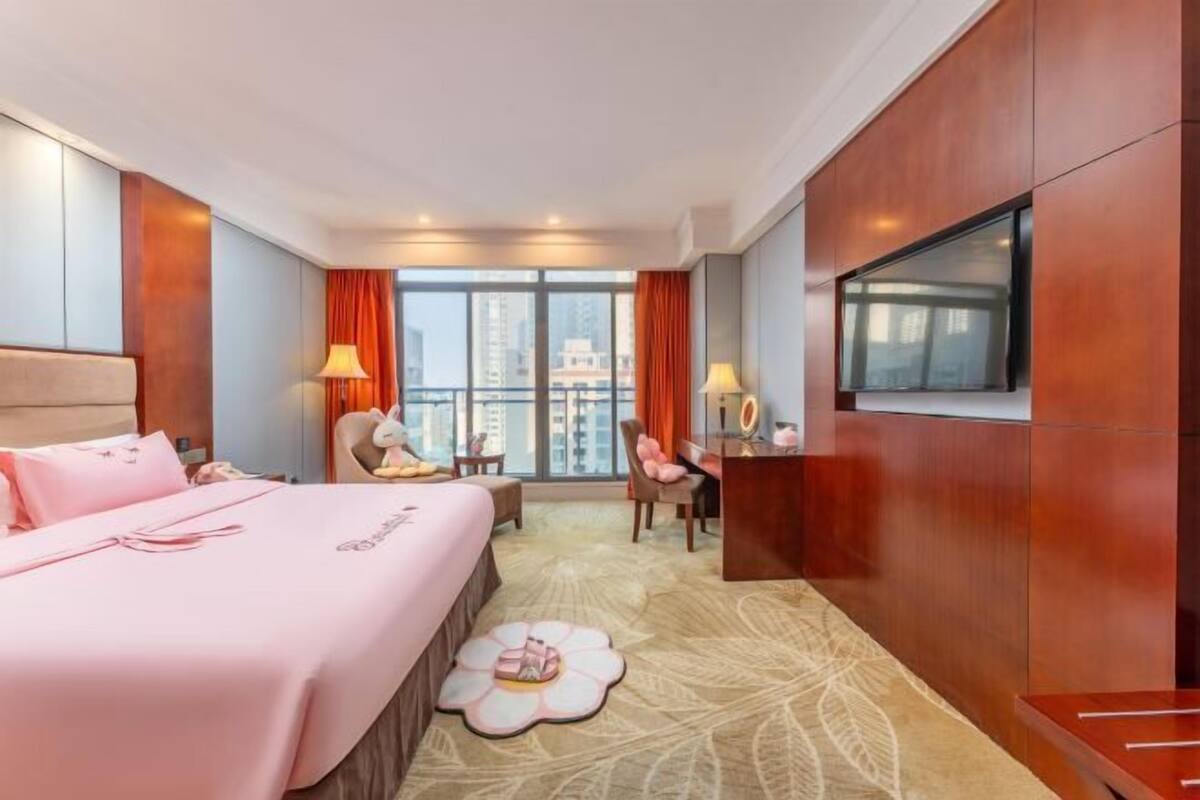 A hotel room with a bed, a desk, a chair, a TV, and a view of the city.