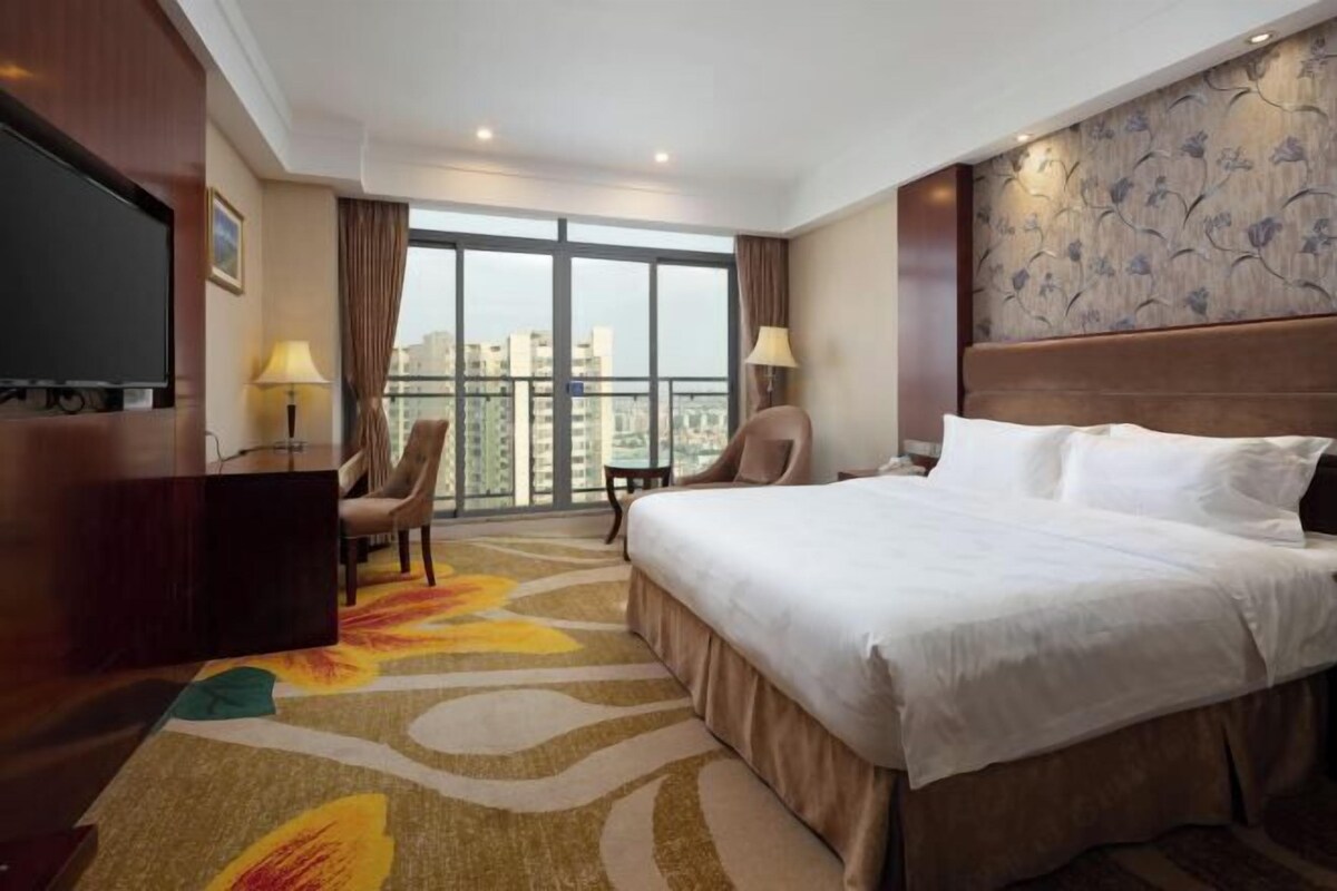 A hotel room with a large bed, a desk, a chair, a TV, and a view of the city through large windows.