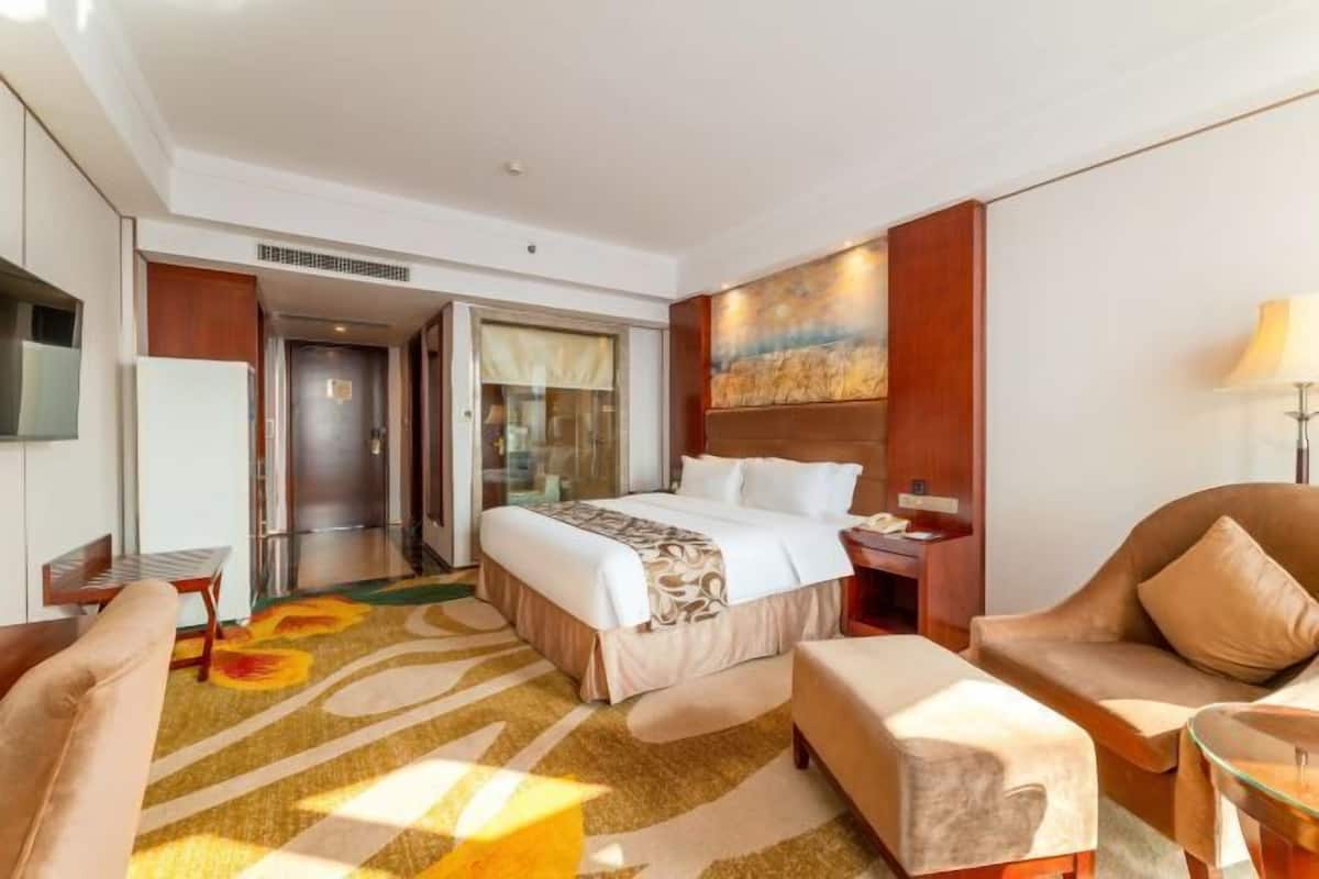 A hotel room with a bed, a sofa, a chair, a glass table, and a painting on the wall.