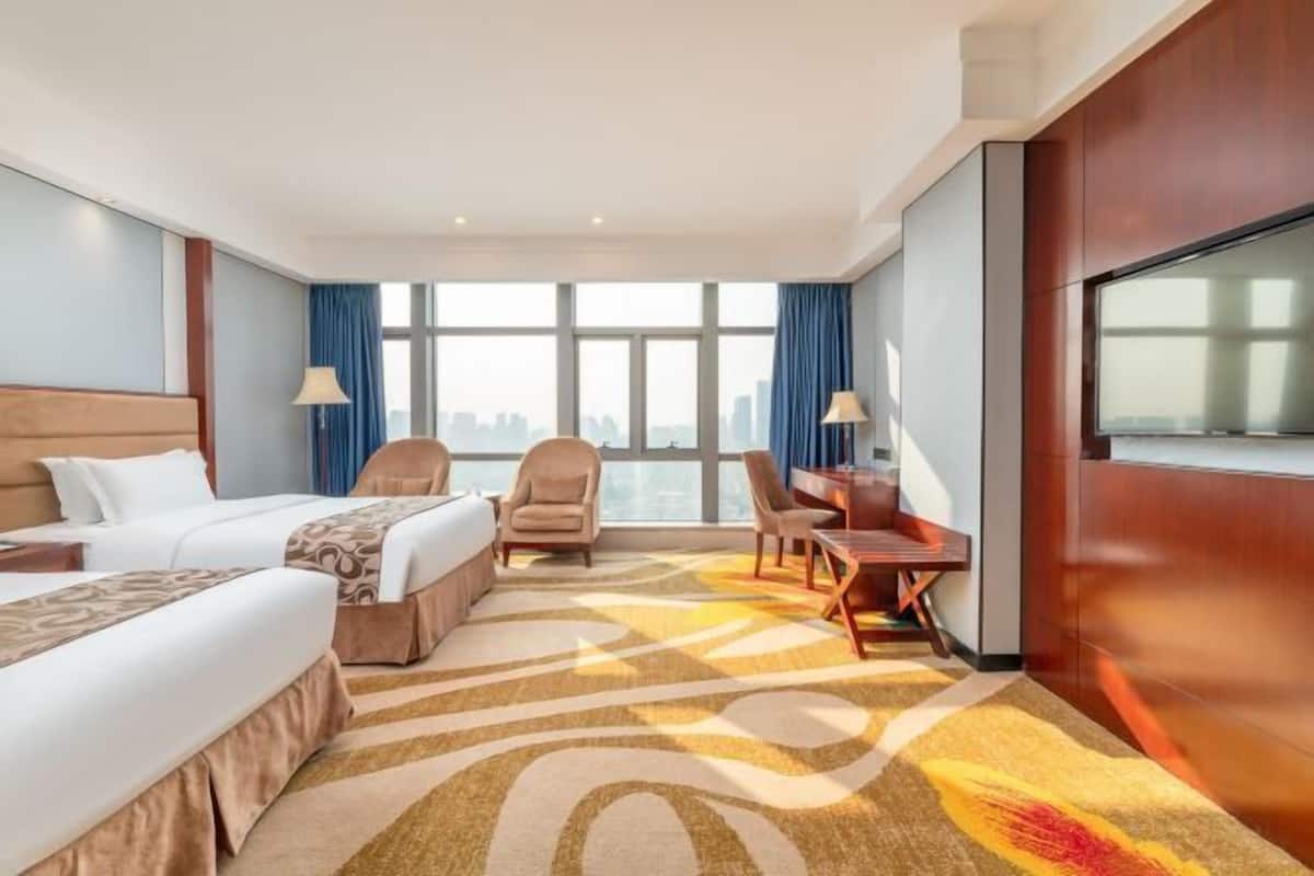 A hotel room with two beds, a desk, a chair, a TV, and a large window with a city view.