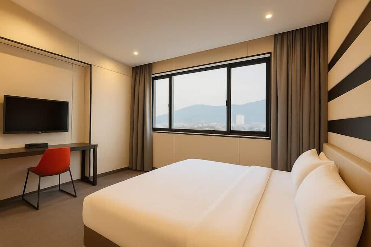 Habitación de hotel con cama, escritorio, silla, televisor y un amplio ventanal con vistas a la ciudad.