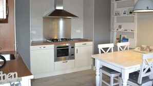 Una cocina moderna con armarios blancos, una campana extractora de acero inoxidable, un horno y una zona de comedor con una mesa de madera y sillas blancas.
