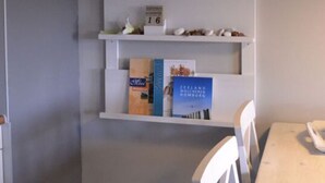 Una habitación con pared blanca, una mesa de madera y sillas blancas. Hay estantes con diversos objetos, incluyendo libros, marcos de fotos y adornos.