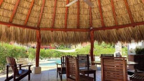 Un espace extérieur abrité, doté d’un toit de chaume, de chaises en bois et d’une piscine en arrière-plan.
