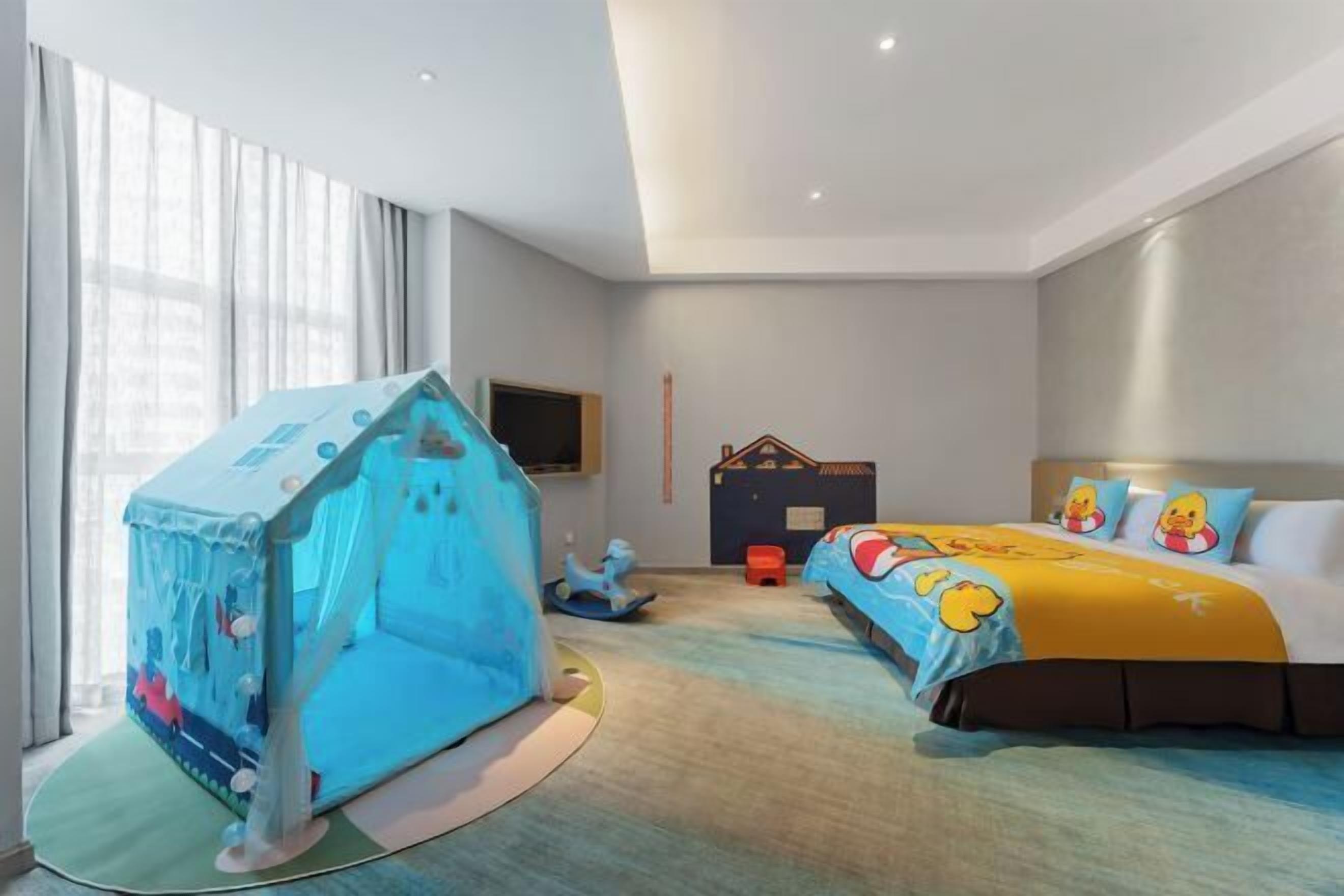 Quarto de hotel com cama, tenda infantil, televisão e janela com cortinas.