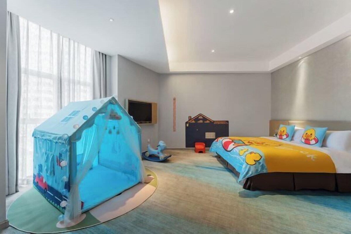 A hotel room with a bed, a play tent, a TV, and a window with curtains.