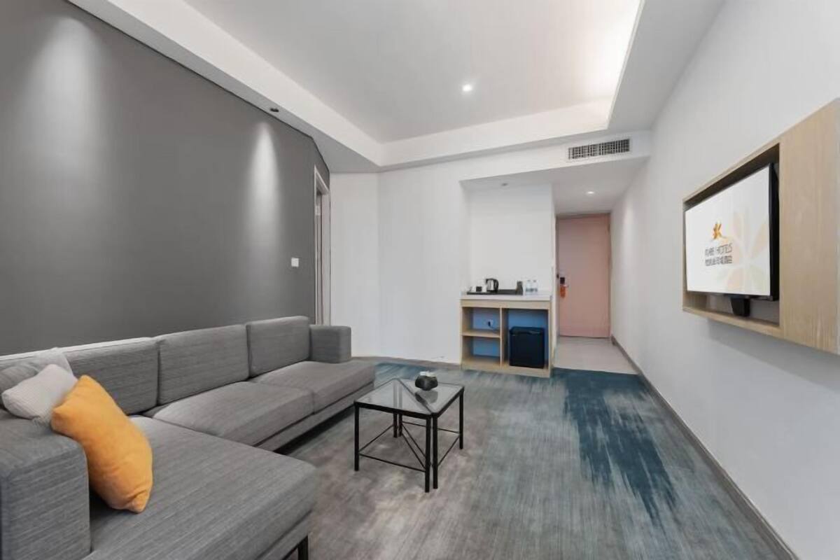 Una habitación de hotel moderna con un sofá gris, una pequeña mesa de vidrio, una kitchenette y un televisor montado en la pared.