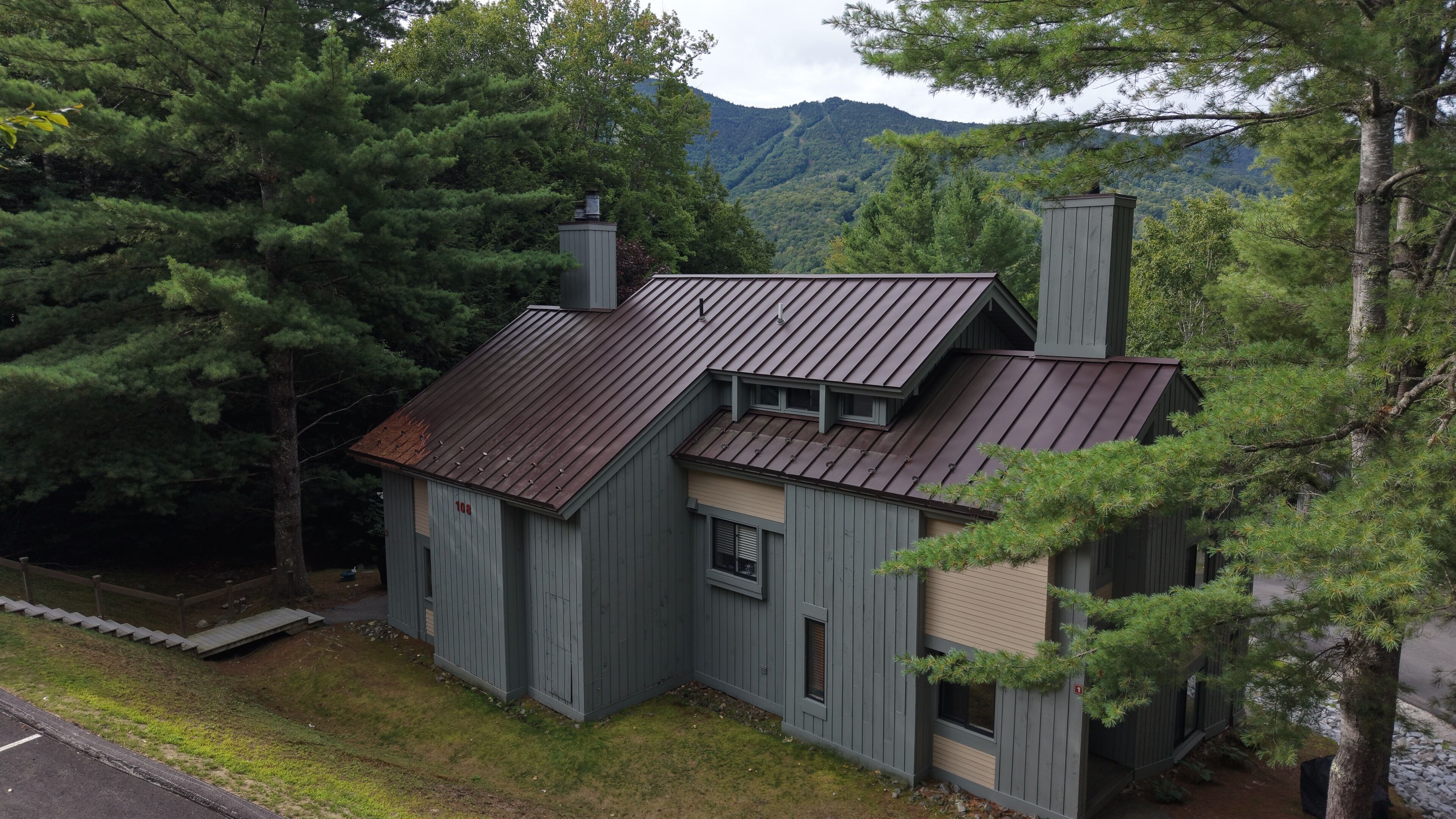 A rustic cabin with a metal roof, surrounded by dense forest and mountains in the background.
