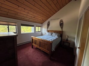 A bedroom with a wooden ceiling, a bed with a wooden headboard, a nightstand, and a window with a view of trees.