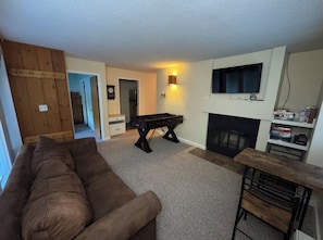 A living room with a brown sofa, a foosball table, a fireplace, and a TV mounted on the wall.
