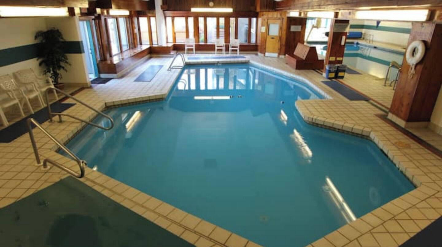 An indoor swimming pool with tiled walls and a wooden floor, surrounded by chairs and a lifebuoy.