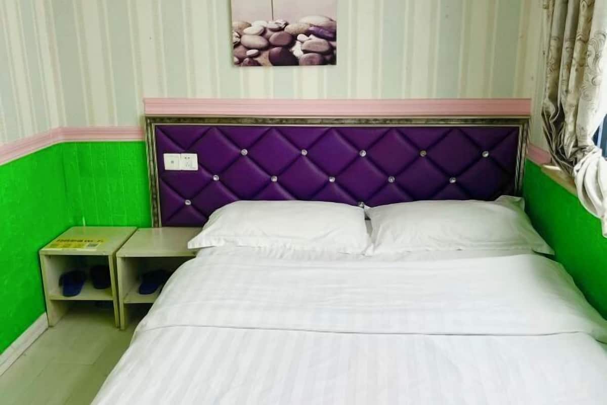 A bed with a purple headboard, white bedding, and a small nightstand with slippers.