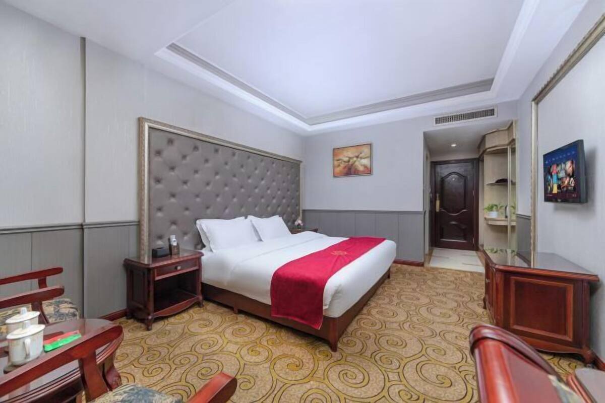 A hotel room with a large bed, a wooden nightstand, a desk with a chair, a red throw blanket, a wall-mounted TV, and a patterned carpet.