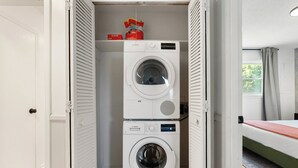 Pieni ja käytännöllinen pyykinhuone, jossa on kaksi päällekkäin asetettua Bosch-pesukonetta, pieni hylly, jossa on pyykinpesuainepussi, ja viereisessä huoneessa sänky, jonka päällä on punainen peitto.
