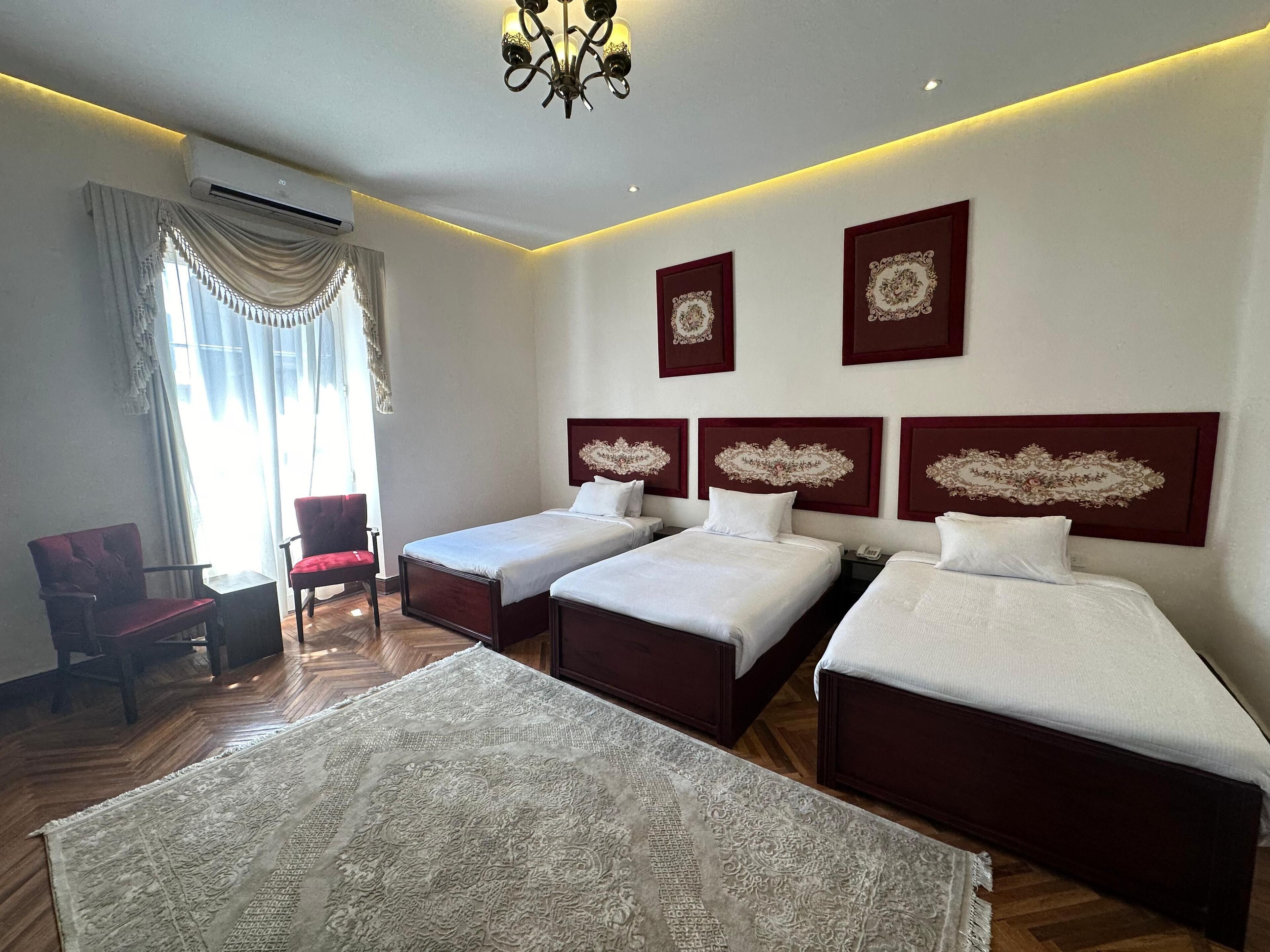 A hotel room with two single beds, a red chair, a large rug, and a chandelier.