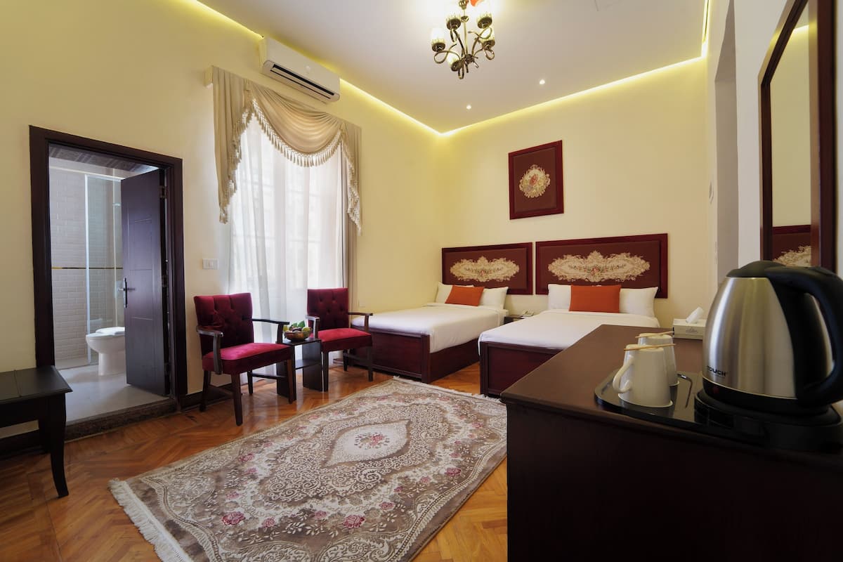Quarto de hotel com uma cama, duas mesas de cabeceira, uma escrivaninha com chaleira, uma cadeira, um tapete e um espelho grande.