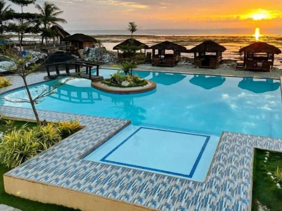 A swimming pool with a view of the sunset over the ocean, surrounded by lounge chairs and umbrellas.