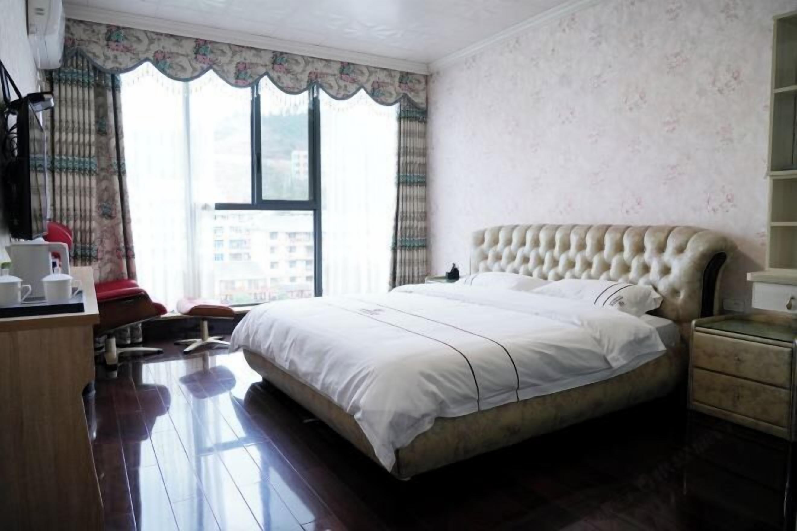 A bedroom with a large bed, a wooden desk, a chair, and a window with curtains.
