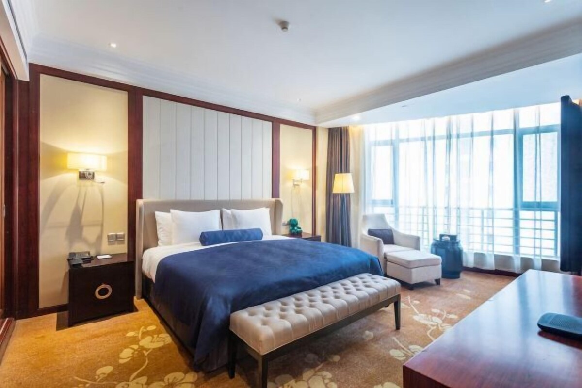 A hotel room with a large bed, a bedside table, a chair, a bench, and a desk.