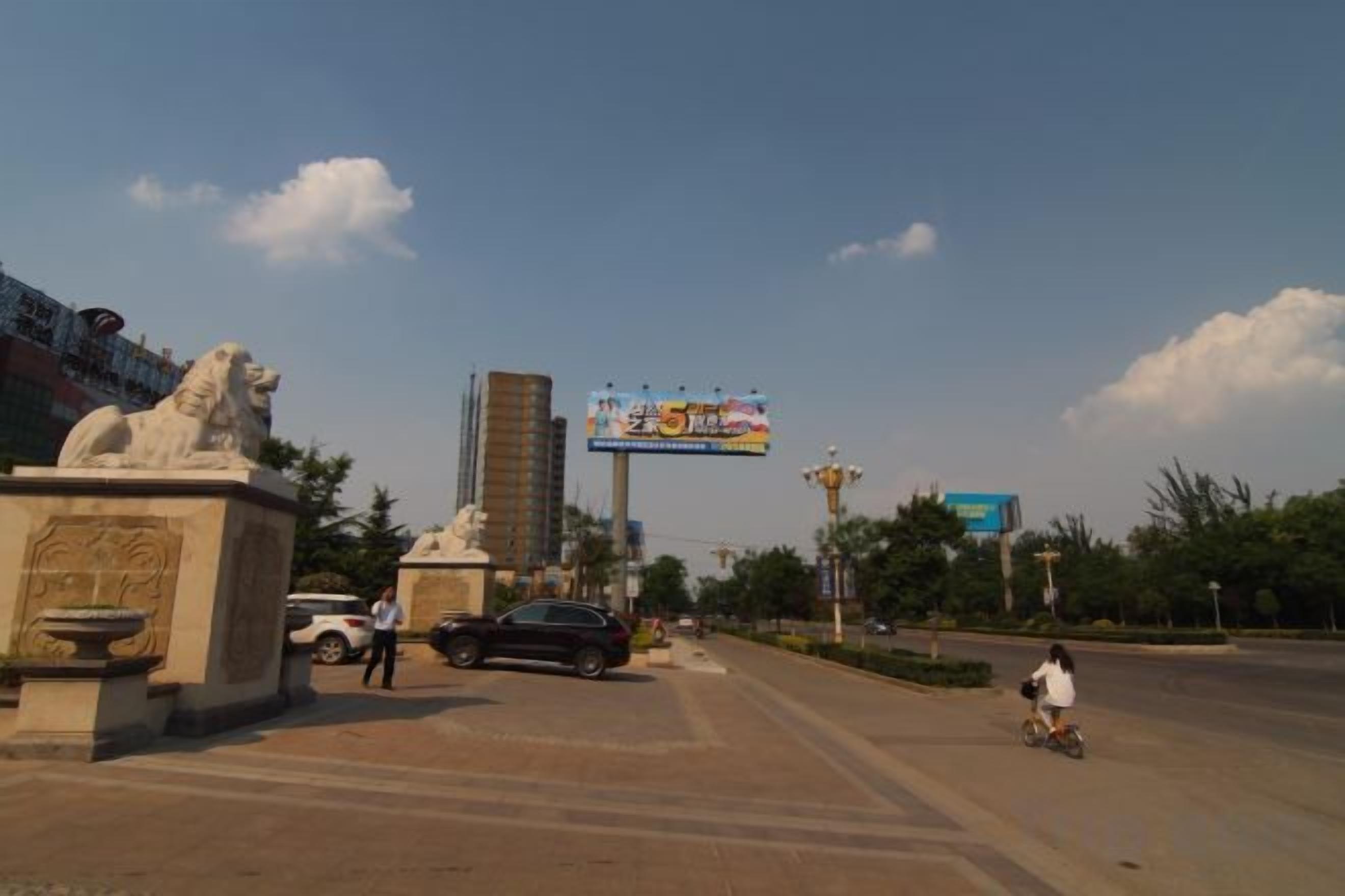 Rua com uma estátua, um outdoor e um edifício ao fundo.