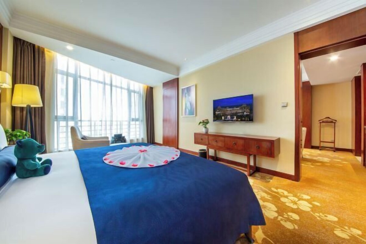 A hotel room with a large bed, a TV on the wall, a desk with a lamp, and a view of the outdoors through large windows.