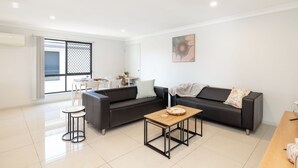 A modern living room with a black sofa, a wooden coffee table, and a dining area with a white table and chairs. - Family Rocklea 3BR | Spacious | Parking | 219D3 (Rocklea)