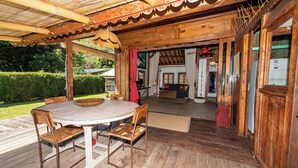 A wooden deck with a round table and chairs, a covered area with a wooden ceiling, and a view into the house. - FARE TEAK with PALAPA and shared pool (Moorea-Maiao)