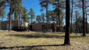 A cabin with a deck and a garage, surrounded by tall pine trees. - Charming 4-bedroom mobile home with WiFi and AC in Timberon (Timberon)