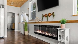 Een moderne woonkamer met een open haard, houten vloeren, een flatscreen televisie aan de muur en een keuken op de achtergrond.