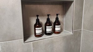 Un baño con azulejos grises, una bañera blanca y tres botellas de jabón corporal, champú y acondicionador en un estante.