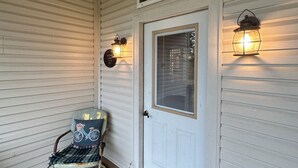 A porch with a wooden bench, a white door with a window, and two wall-mounted lanterns.