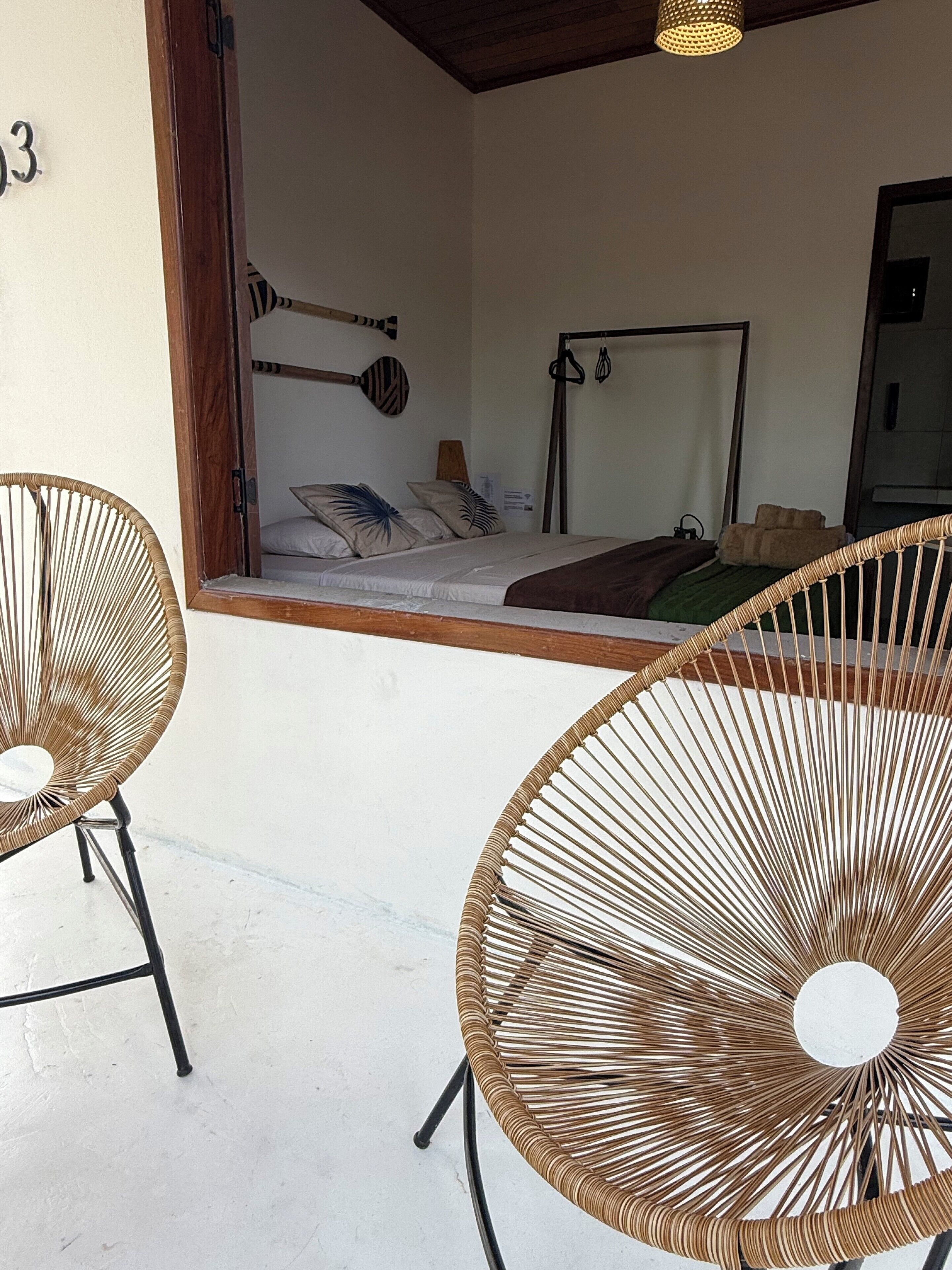 Una habitación con dos sillones de mimbre, una cama con cabecera, un espejo fijado a la pared y una lámpara en el techo.
