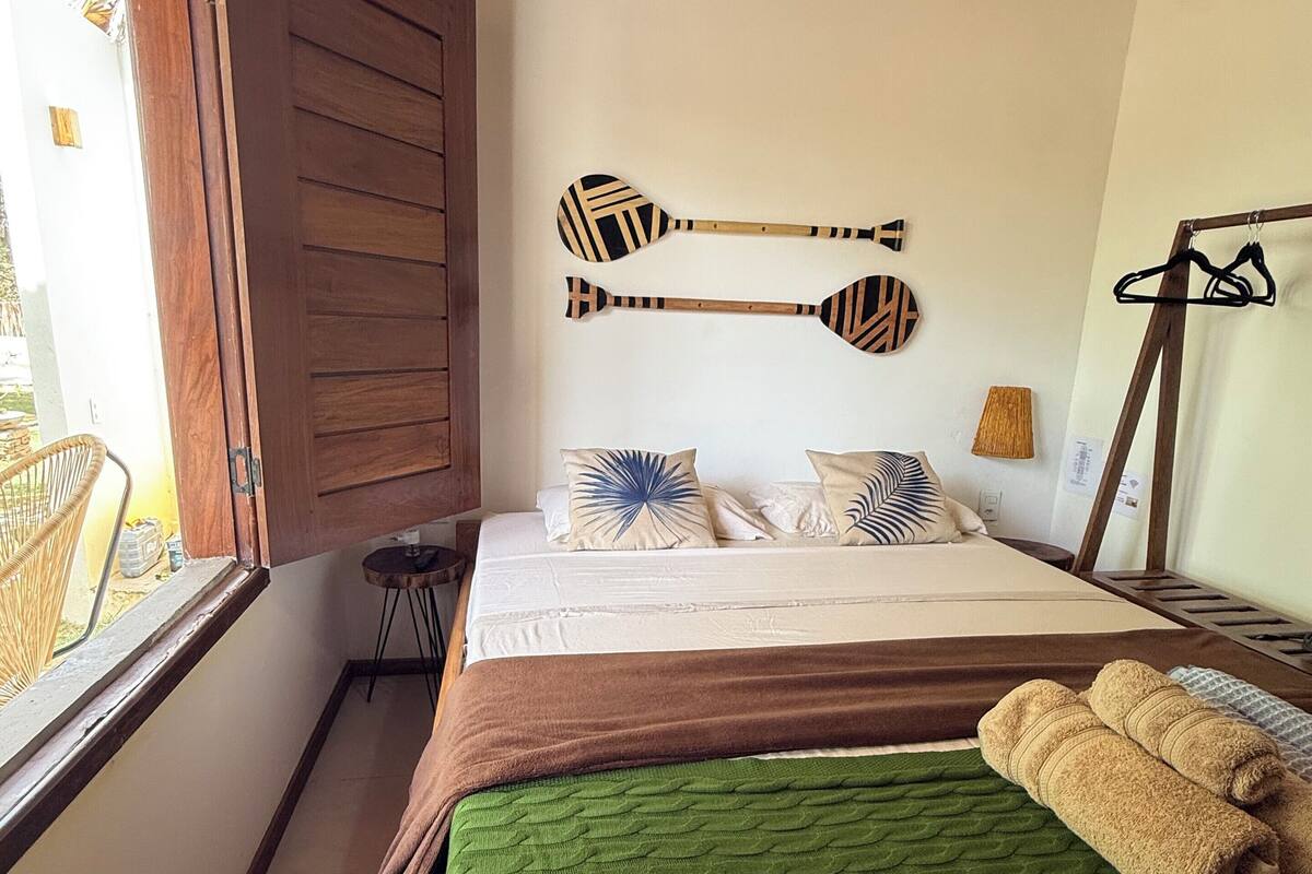Una camera da letto con un letto di legno, una coperta verde e due asciugamani. Si notano due decorazioni a forma di pagaia appese al muro e un tavolino con una lampada.