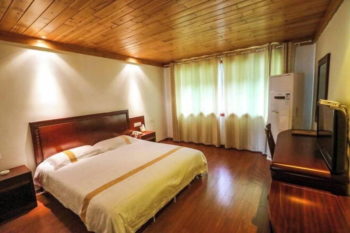 A hotel room with a wooden ceiling, a bed with white bedding, a nightstand, a desk with a chair, and a window with curtains.