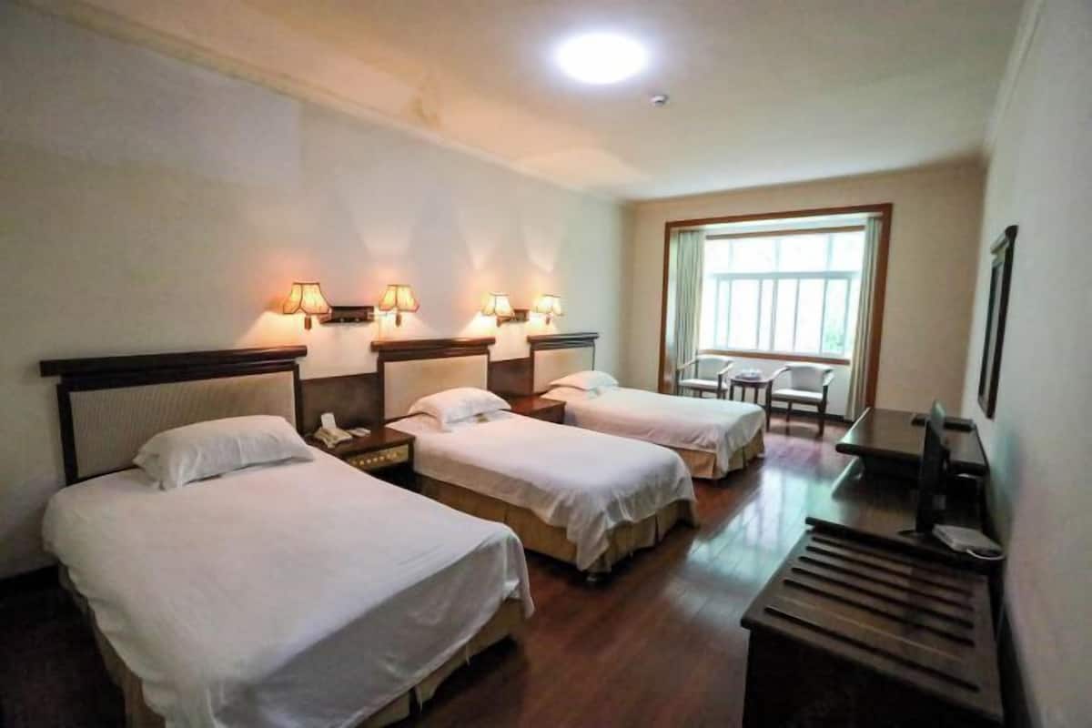 A hotel room with three beds, bedside lamps, a desk with a chair, a window, and a desk lamp.