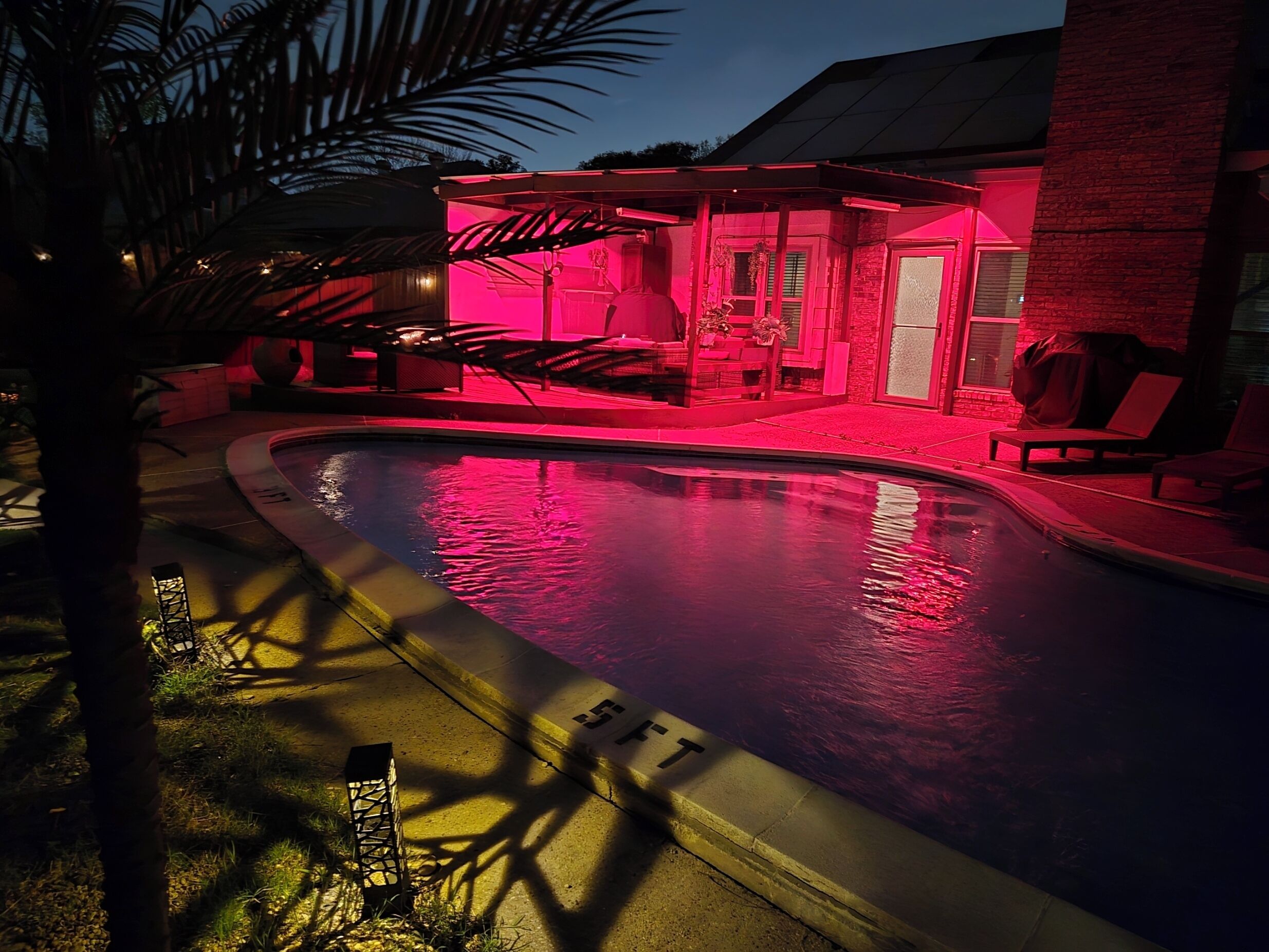 A swimming pool with lounge chairs and a covered seating area, surrounded by a brick building.