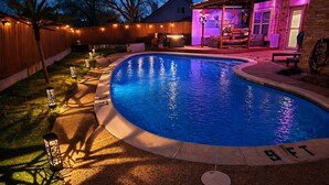 A swimming pool with a wooden deck, a covered patio area, and a fenced backyard.