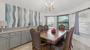 A dining room with a wooden table, six chairs, a chandelier, and a large abstract painting on the wall.