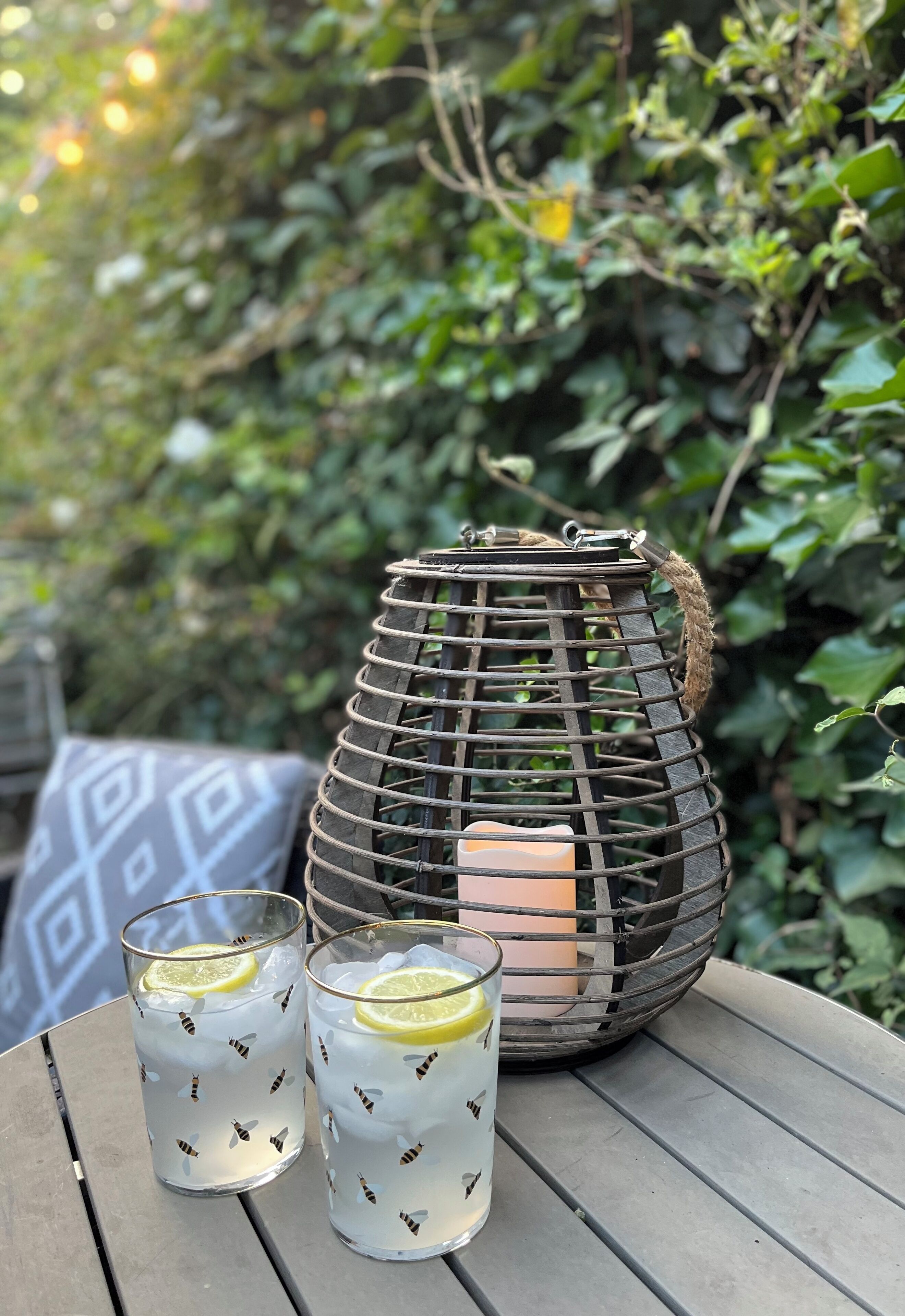 Twee glazen met citroenpartjes op een tafel, een kaars in een metalen houder, en een gestreept kussen in een tuin.