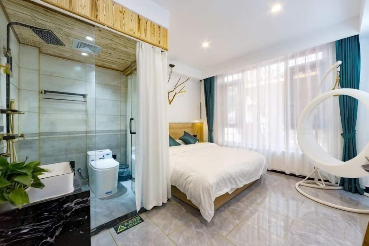 Moderni hotellihuone, jossa on lasiseinällinen suihku, valkoisella verholla varustettu sänky ja riipputuoli.