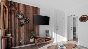 A modern living room with a wooden accent wall, a flat-screen TV, a wooden coffee table, a grey sofa, and a white door leading to another room. - Private Garden Retreat - King Bed - mins TMH + Downtown + FSU! (Tallahassee)