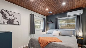A bedroom with a large bed, two windows with curtains, a wall-mounted air conditioner, and a wooden ceiling. - Private Garden Retreat - King Bed - mins TMH + Downtown + FSU! (Tallahassee)
