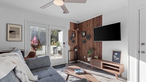 A modern living room with a grey sofa, a wooden coffee table, a flat-screen TV mounted on a wooden wall, and a ceiling fan. - Private Garden Retreat - King Bed - mins TMH + Downtown + FSU! (Tallahassee)